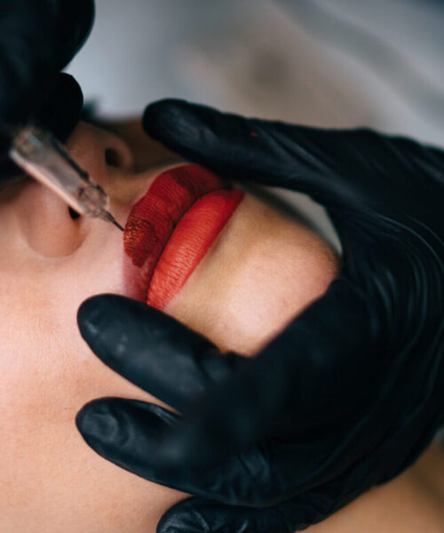 Partial image of beautician doing permanent makeup lips procedure with needle tool of caucasian woman client in beauty salon. Professional female master wearing gloves. Beauty concept